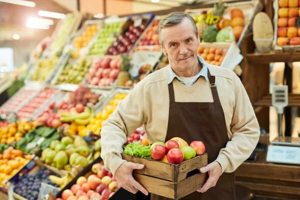 Waist up portrait of smiling senior man holding box of apples while selling fruits at farmers market